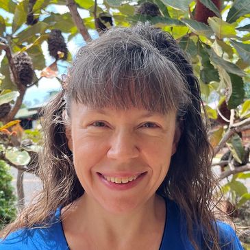 Smiling woman with long hair in blue shirt outdoors near plants.
