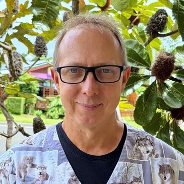 A man with glasses stands outdoors among lush foliage, wearing a shirt with animal prints.