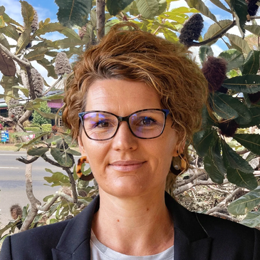Woman with glasses and curly hair standing outdoors near leafy plants.