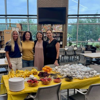 Four women standing behind a table with food in a library.