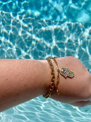 Wrist adorned with two gold bracelets, one featuring a colorful Hamsa charm, against a shimmering pool backdrop.