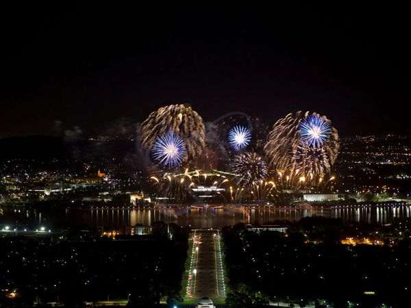 Nighttime fireworks light up the sky over a cityscape with a river and illuminated streets.