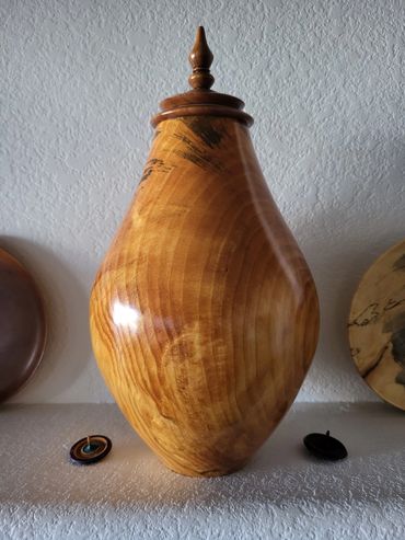 Polished wooden vase with a pointed lid on a textured white surface.