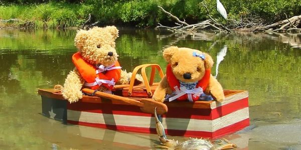 Two teddy bears in a tiny wooden boat in the water. The girl teddy bear is looking over the edge of