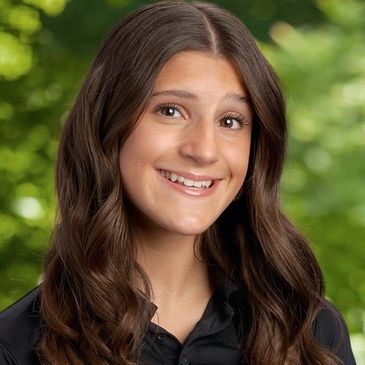 Smiling young woman with long brunette hair against a green leafy background.