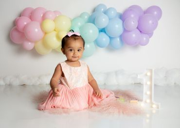 Girl sits on a photography set decorated with pastel balloons for her first birthday photoshoot.