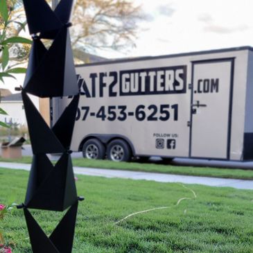 Black geometric rain chain hanging outdoors with a gutter service trailer in background.