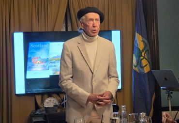 An elderly man in a beret speaks in front of a Scotland presentation.