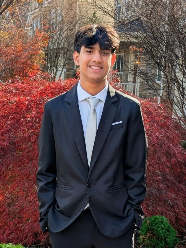 Young man in a black suit standing outdoors with autumn foliage behind him.