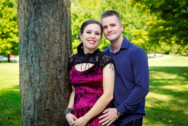 A couple posing in a sunny park, with the woman leaning against a tree and smiling brightly.