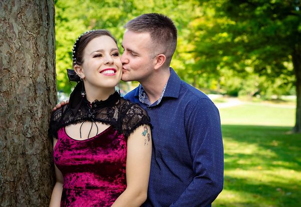 A couple in a park, with the man kissing the smiling woman on the cheek as she leans against a tree.