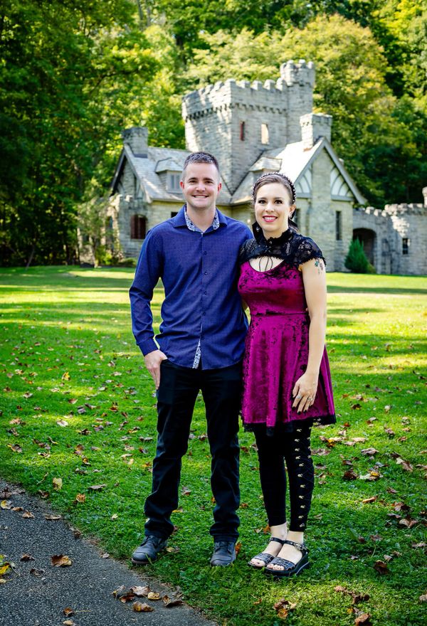A couple posing together in a grassy park with a stone castle-like building in the background.