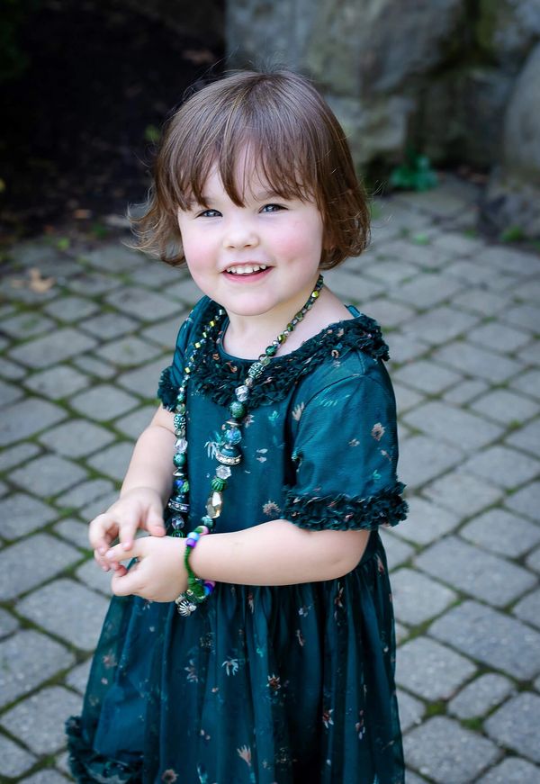 A smiling toddler in a green dress and beaded necklace standing on a cobblestone path.