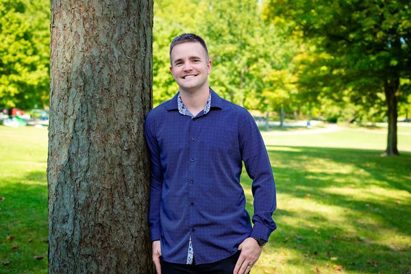 A man in a blue button-up shirt leaning against a tree in a sunny park setting.