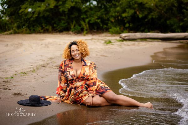 Smiling woman in a patterned dress sitting on a beach with waves touching her feet.