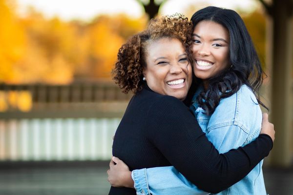 Mother and daughter embrace and smile at the camera in an outdoor setting.
