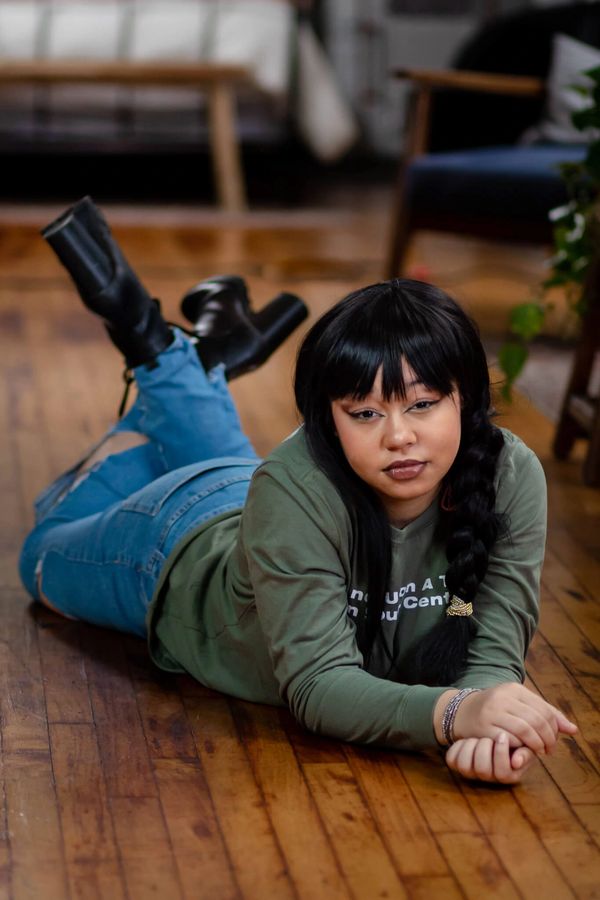 A woman with dark hair lies on a wooden floor, wearing jeans, a green sweater, and black boots.