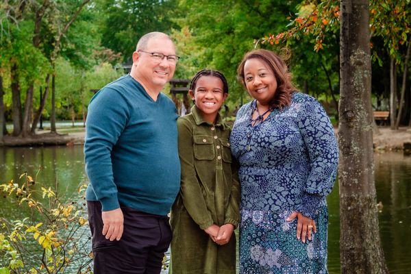 Smiling family of three posing by a pond with trees in the background.