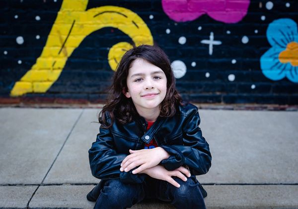 A smiling boy in a black leather jacket sits in front of a colorful mural.