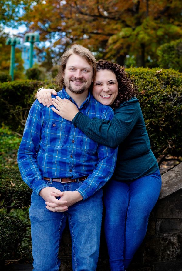 A smiling couple sitting together on a stone bench in a park with autumn foliage in the background.