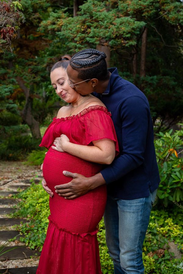 Man kissing pregnant woman's cheek in a garden while holding her pregnant belly.