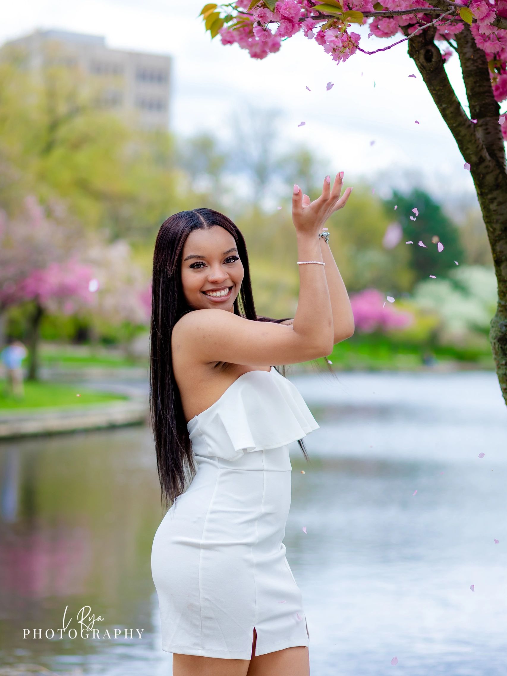 High school senior girl in white dress smiling while catching cherry blossom petal falling off a tre