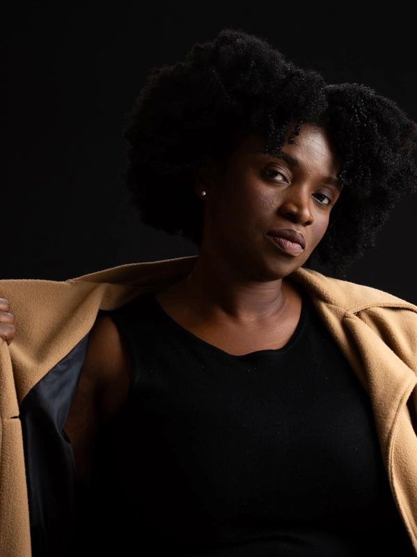 Confident woman in black top and tan coat against dark background.