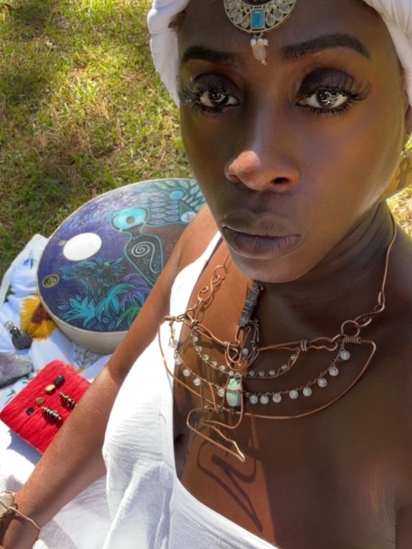 Woman in white dress with ornate jewelry and drum outdoors.