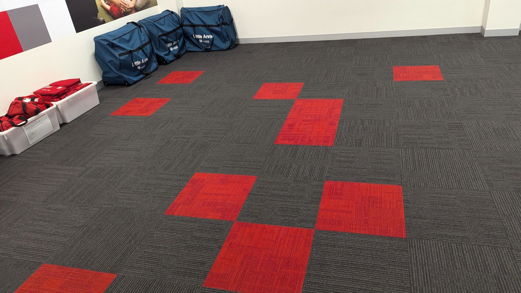 Room with gray and red carpet tiles and blue bags labeled Little Anne.