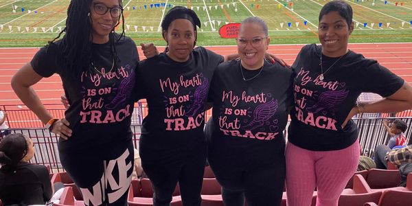 Four women wearing matching 'My heart is on that track' shirts at a stadium.
