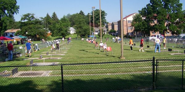 People playing horseshoes in a grassy park area with houses and trees in the background.