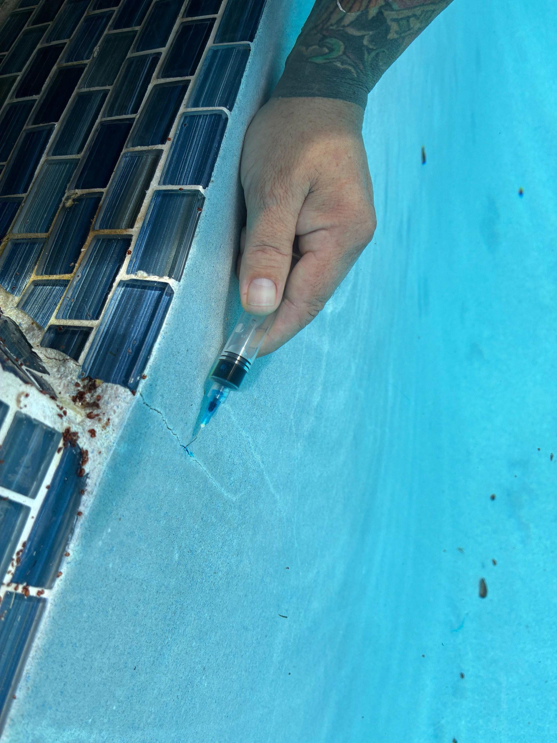 A hand with a tattoo holds a syringe near a cracked pool edge.