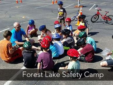 Students in bike helmets sit in a group listening to an adult. Children's' bikes parked in the back.