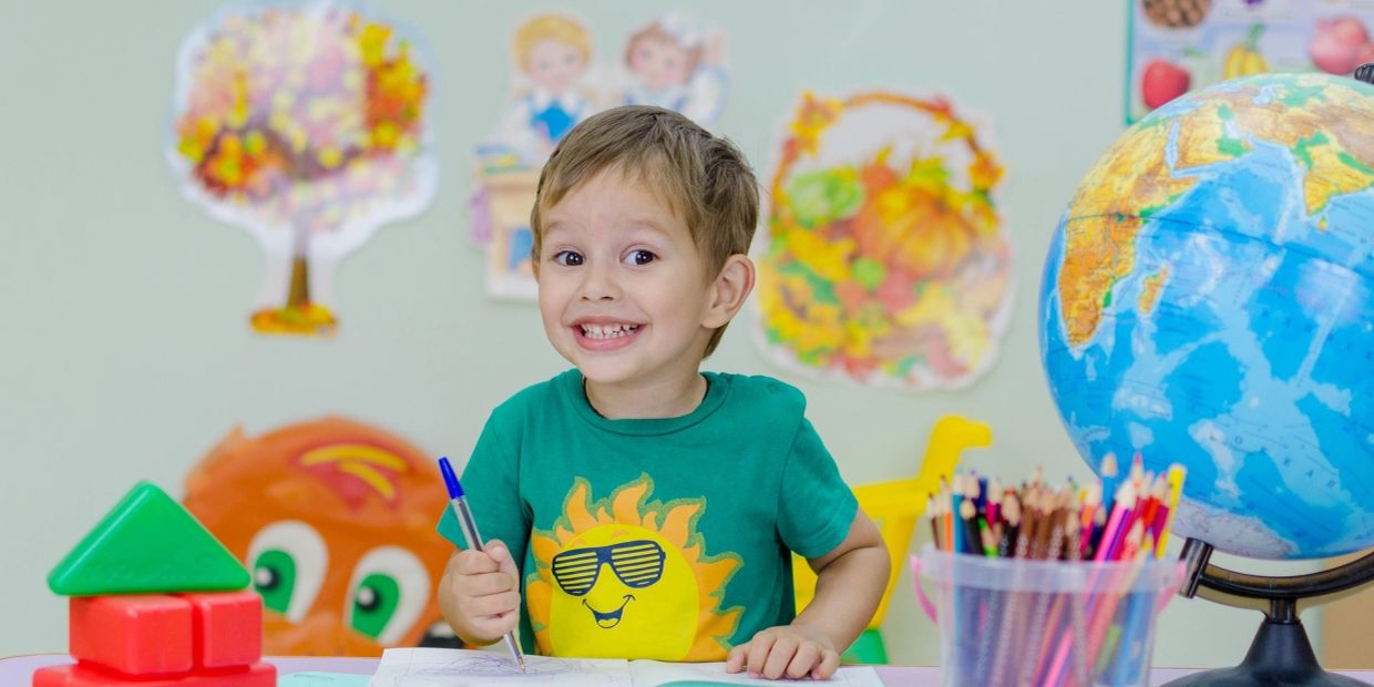 Smiling boy drawing at a colorful classroom table.
