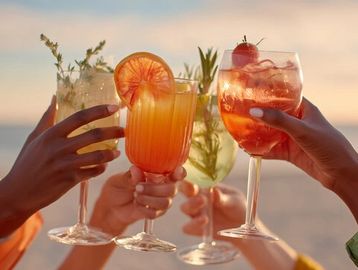 Hands clinking colorful cocktails against a sunset backdrop.