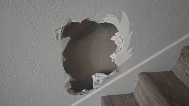 Hole in the stairwell drywall before repair, beside the wooden stairs and white trim.