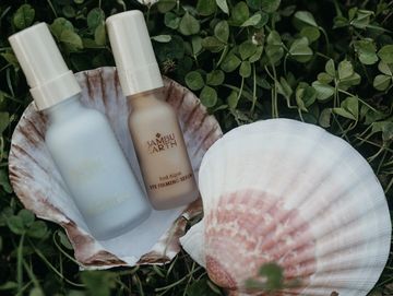 Two cosmetic bottles in a seashell on greenery.