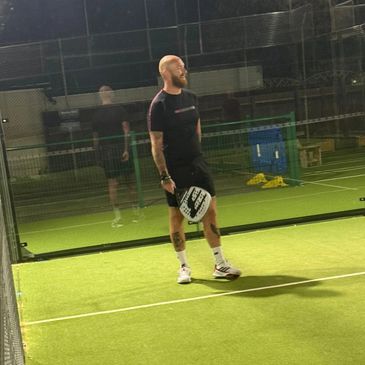 Man standing on a lit paddle tennis court at night, holding a paddle.