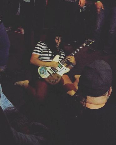 Hazy photo of a woman playing guitar on her knees on the floor surrounded by  a crowd of people. She