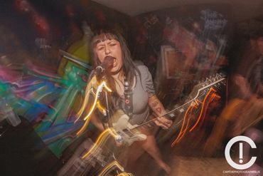 Blurry photo of a woman singing into a microphone and playing guitar. She is barefoot and wearing a 