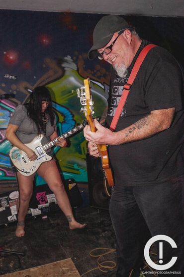 A smiling man and a woman play guitars in front of a colorful graffiti wall. The woman is barefoot. 
