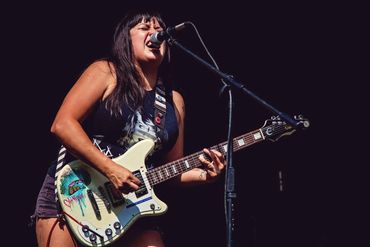 A woman plays guitar and singing into a microphone on an outside stage on a bright sunny day. She is