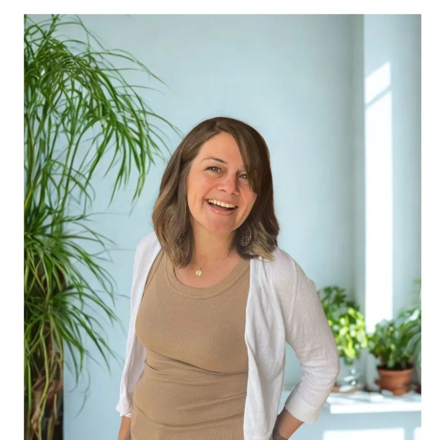 A smiling woman stands indoors with plants in the background.