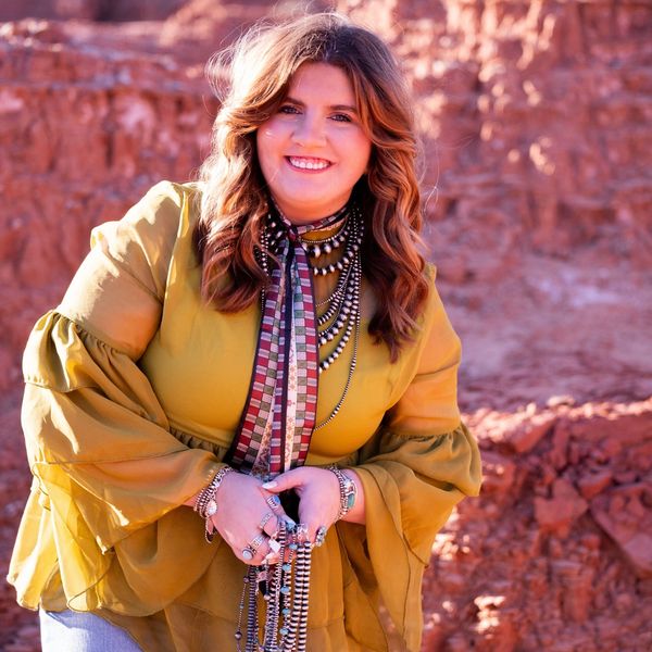 A smiling woman in a yellow blouse with layered necklaces poses outdoors against a rocky background.