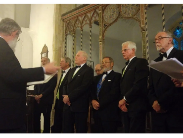 A men's choir dressed in tuxedos performs under a conductor in a historic church.