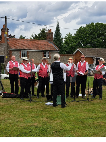 A group of men in red vests singing outdoors with a conductor.