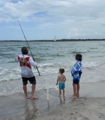 Three people on the beach, one holding a fishing rod, looking at the ocean.