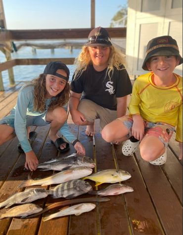 Three kids proudly display their freshly caught fish on a wooden dock near the water.