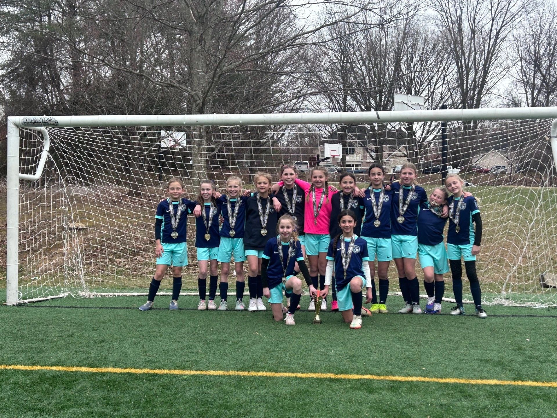 Youth soccer team posing with medals and a trophy on a soccer field.