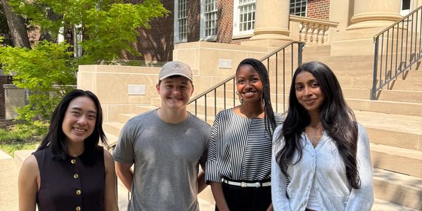 Four young adults smiling outdoors in front of a building with stairs.